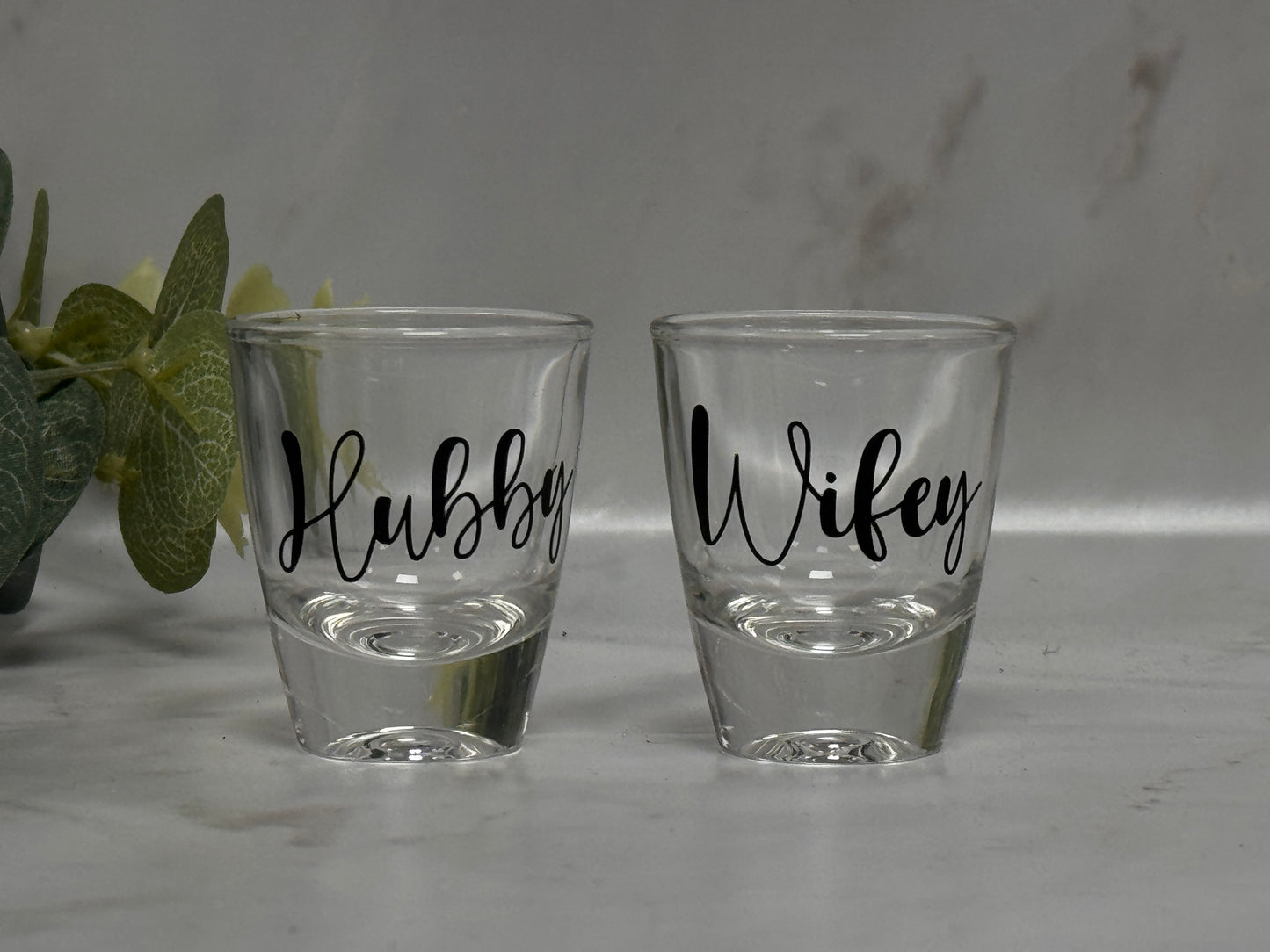 Two shot glasses labeled 'Hubby' and 'Wifey' on a light surface.