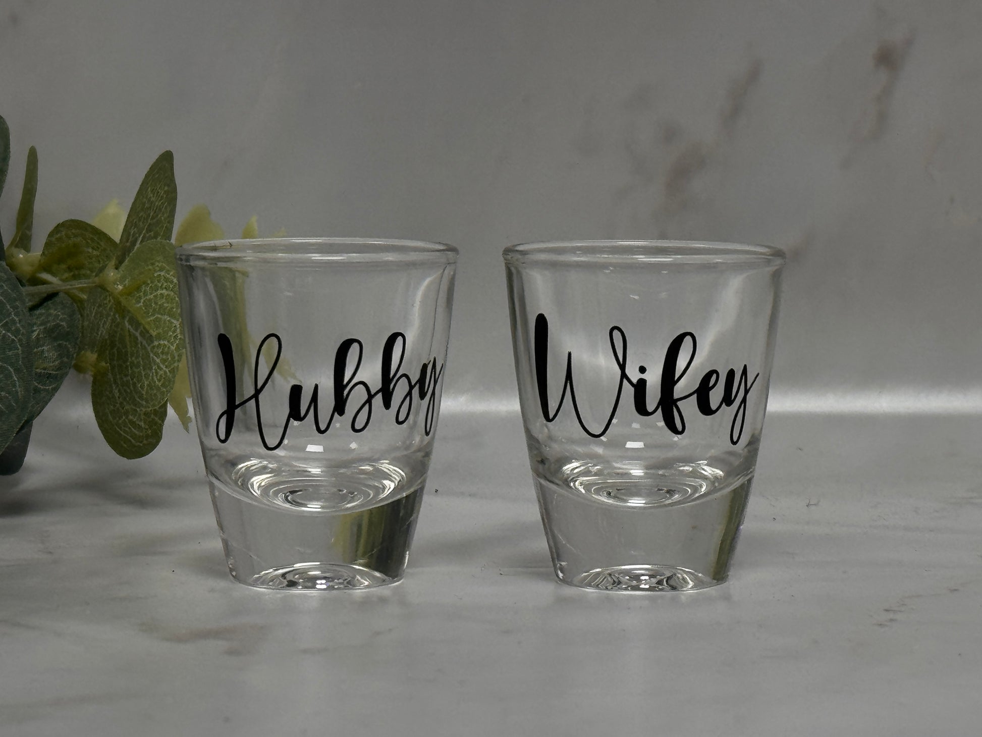 Two shot glasses labeled 'Hubby' and 'Wifey' on a light surface.