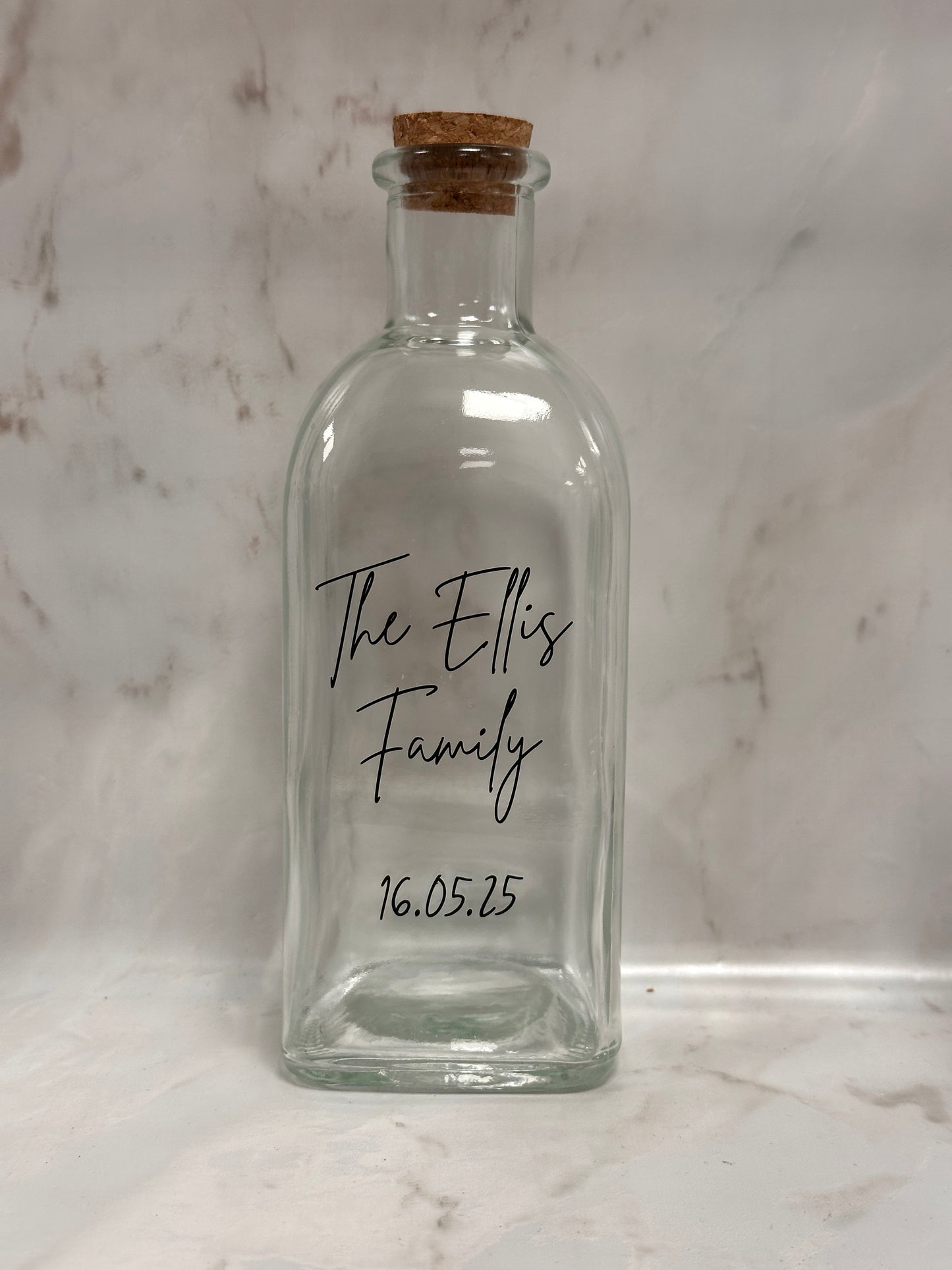 Clear glass bottle with cork stopper on a marble surface, featuring 'The Ellis Family' and a date.